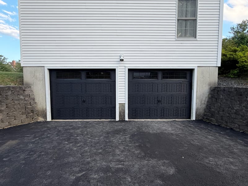 Two new black carriage-style garage doors with windows and decorative handles between stone retaining walls
