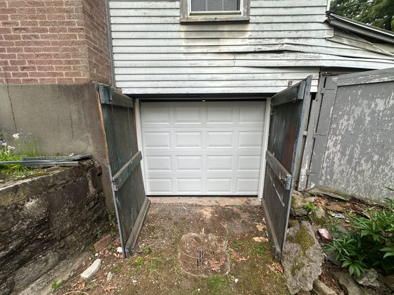 New white sectional garage door in a tight opening under a house, replacing old wooden swing doors